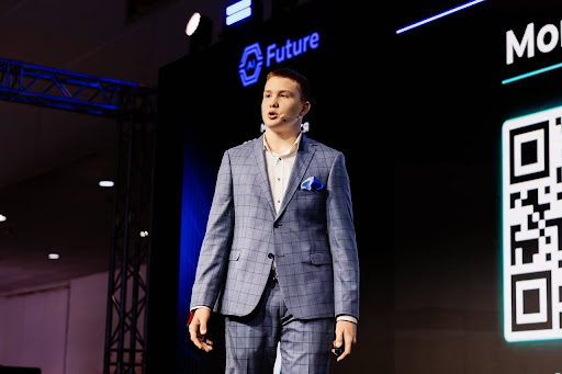 Nikita speaking on stage at a blockchain forum, wearing a sharp suit, illuminated by neon blue lights in a premium tech conference setting.